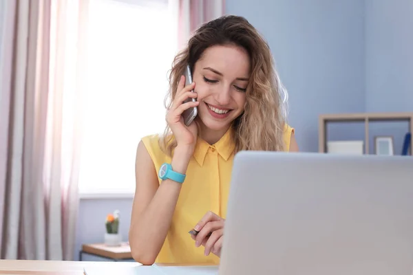 Jonge vrouw die met een mobiele telefoon praat terwijl ze met een laptop aan het bureau werkt. Binnenlandse Zaken — Stockfoto