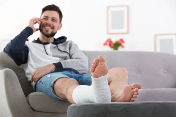 Homem com a perna quebrada no elenco usando telefone celular enquanto sentado no sofá em casa — Fotografia de Stock