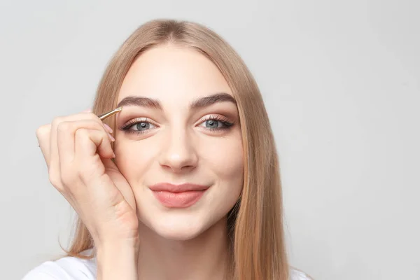 Jonge vrouw plukken de wenkbrauwen met pincet op lichte achtergrond — Stockfoto