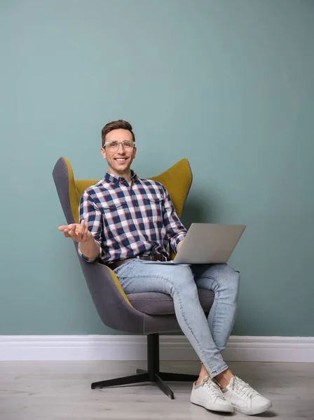 Jeune blogueur avec ordinateur portable assis dans un fauteuil contre un mur de couleur — Photo