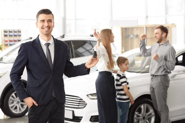 Vendedor com chave e jovem família no salão de carro — Fotografia de Stock