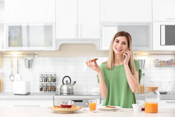 Hermosa mujer comiendo sabroso pan tostado con mermelada mientras habla en el teléfono móvil en la mesa en la cocina —  Fotos de Stock