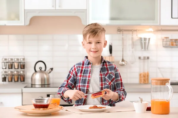 Cute little boy spreading jam onto tasty toasted bread at table in kitchen
