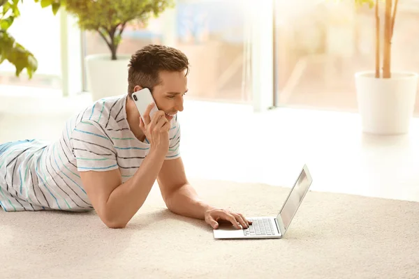 Jovem bonito com telefone celular e laptop no tapete acolhedor em casa — Fotografia de Stock