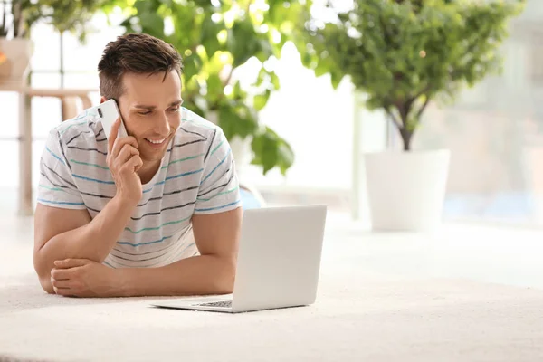 Jovem bonito com telefone celular e laptop no tapete acolhedor em casa — Fotografia de Stock