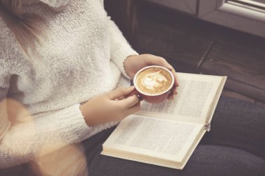 Elinde kahve fincanı olan bir kadın evde kitap okuyor.