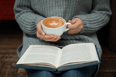 Elinde kahve fincanı olan bir kadın evde kitap okuyor.