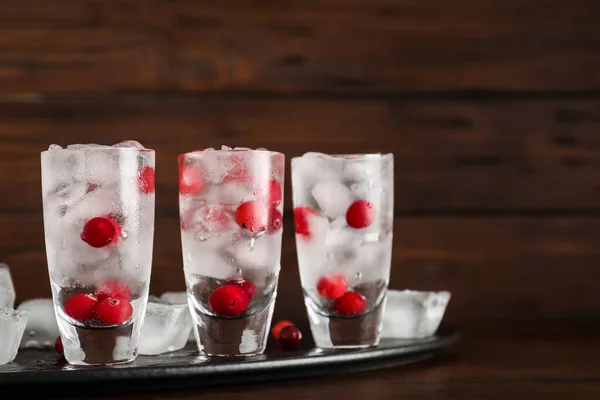 Shot glasses with vodka, ice and cranberries on table against wooden background — Stock Photo, Image