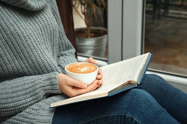 Mujer con taza de libro de lectura de café cerca de la ventana en el interior, primer plano — Foto de Stock