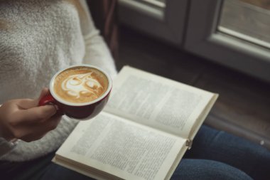 Elinde kahve fincanı olan kadın pencerenin yanında kitap okuyor, yakın plan.