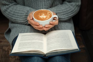 Elinde kahve fincanı olan bir kadın evde kitap okuyor.