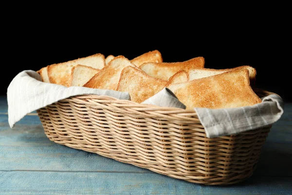浅蓝色木制桌子上的篮子里的烤面包片，背景为黑色 — 图库照片