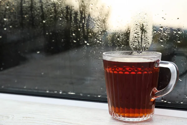 Szklana filiżanka gorącej herbaty na białym drewnianym parapecie, zbliżenie. Pogoda deszczowa — Zdjęcie stockowe