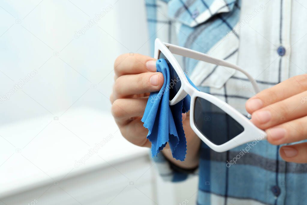 Woman wiping sunglasses with microfiber cleaning cloth indoors, closeup
