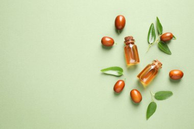 Glass bottles with jojoba oil and seeds on green background, flat lay. Space for text clipart