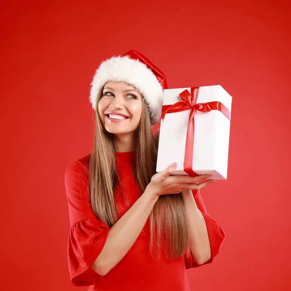 Счастливая молодая женщина в шляпе Санты с рождественским подарком на красном фоне — стоковое фото
