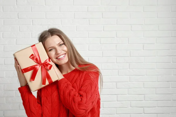 Mujer joven feliz con regalo de Navidad cerca de la pared de ladrillo blanco. Espacio para texto —  Fotos de Stock