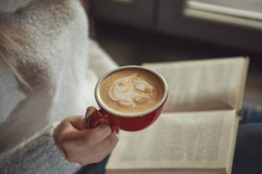 Elinde kahve fincanı olan kadın pencerenin yanında kitap okuyor, yakın plan.