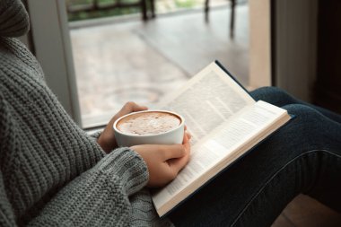 Elinde kahve fincanı olan kadın pencerenin yanında kitap okuyor, yakın plan.