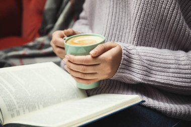 Elinde kahve fincanı olan bir kadın evde kitap okuyor.