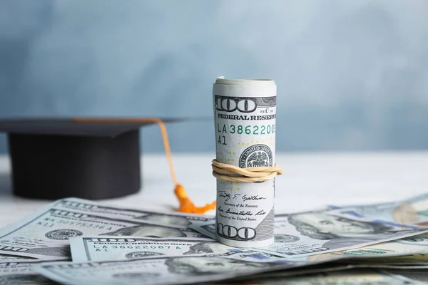 Dollar banknotes and student graduation hat on white wooden table against blue background. Tuition fees concept