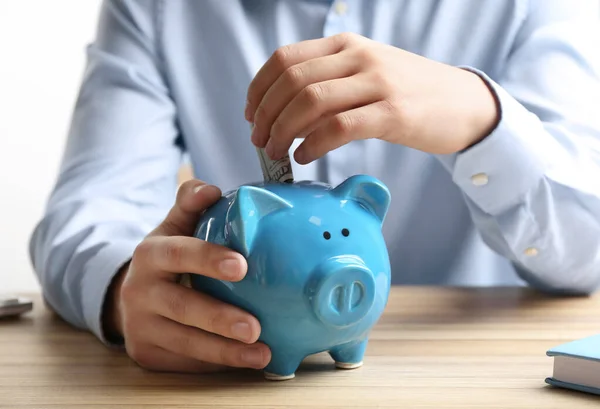 Hombre poniendo billete de dólar en alcancía en la mesa de madera en el interior, primer plano. Ahorro de dinero — Foto de Stock