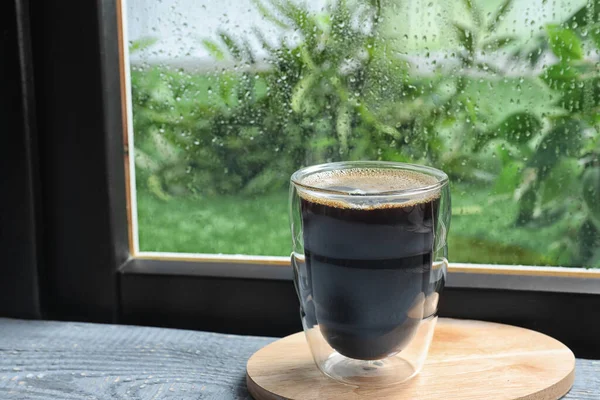 Glass of coffee on wooden windowsill, space for text. Rainy weather