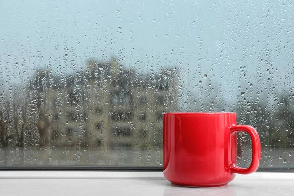 Taza de bebida caliente en el alféizar de la ventana, espacio para el texto. Clima lluvioso — Foto de Stock