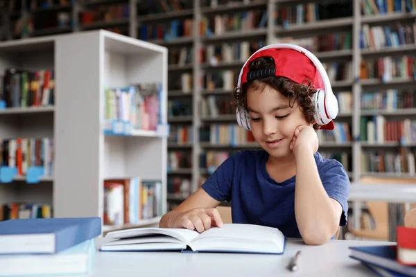 Menino bonito com fones de ouvido lendo livros à mesa na biblioteca — Fotografia de Stock