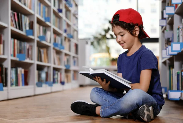 Lindo niño leyendo libro en el suelo en la biblioteca — Foto de Stock