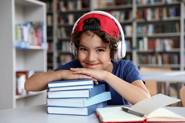 Lindo niño con auriculares y libros en la mesa en la sala de lectura de la biblioteca — Foto de Stock