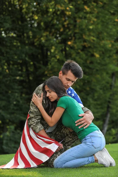 Homem de uniforme militar com bandeira americana abraçando sua esposa no parque verde — Fotografia de Stock