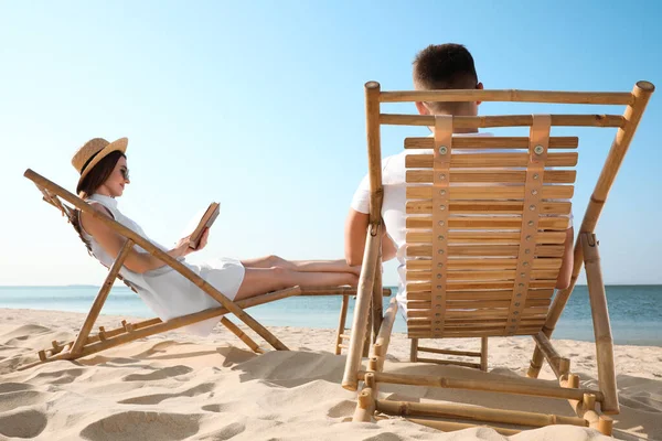 Νεαρή γυναίκα διαβάζει βιβλίο και ο άνθρωπος χαλαρώνοντας σε ξαπλώστρες στην παραλία κοντά στη θάλασσα — Φωτογραφία Αρχείου