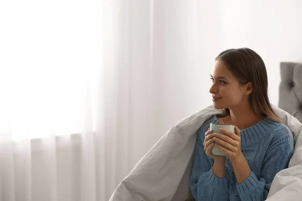 Молодая женщина в теплом свитере с горячим напитком на кровати дома. Пространство для текста — стоковое фото