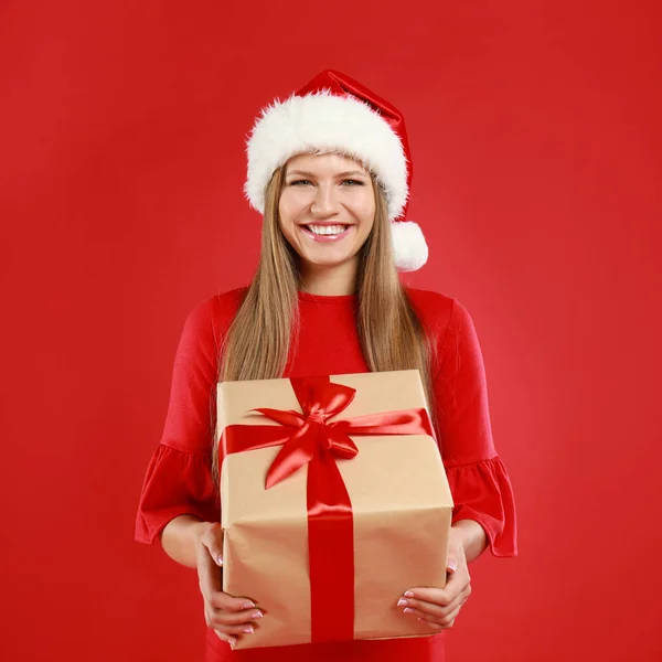 Ευτυχισμένη νεαρή γυναίκα σε Σάντα καπέλο με χριστουγεννιάτικο δώρο σε κόκκινο φόντο — Φωτογραφία Αρχείου