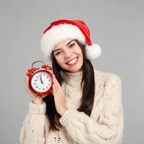 Ευτυχισμένη νεαρή γυναίκα σε Σάντα καπέλο με ξυπνητήρι σε γκρι φόντο. Χριστούγεννα — Φωτογραφία Αρχείου