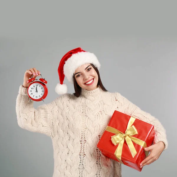 Joyeux jeune femme dans le chapeau de Père Noël avec cadeau de Noël et réveil sur fond gris — Photo