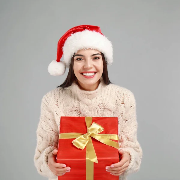Счастливая молодая женщина в шляпе Санты с рождественским подарком на сером фоне — стоковое фото