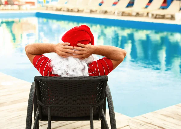 Аутентичный Санта-Клаус отдыхает на кресле у бассейна на курорте, вид сзади — стоковое фото