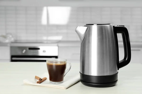 Современный электрический чайник, чашка кофе и коричные палочки на белом деревянном столе на кухне — стоковое фото