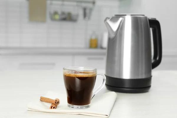 Modern electric kettle, cup of coffee and cinnamon sticks on white wooden table in kitchen — ストック写真