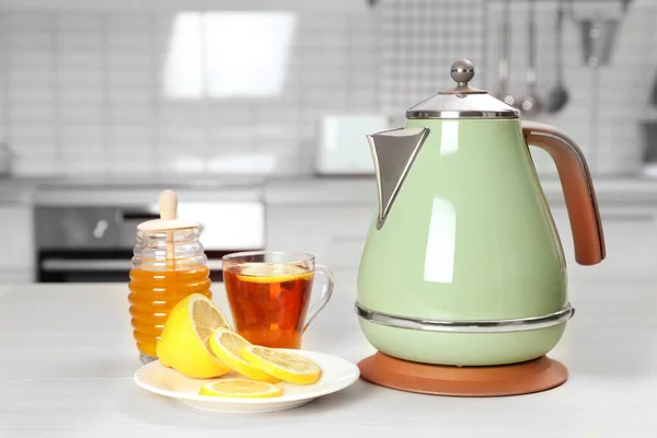 Современный электрический чайник, чашка чая и лимон на белом деревянном столе на кухне — стоковое фото