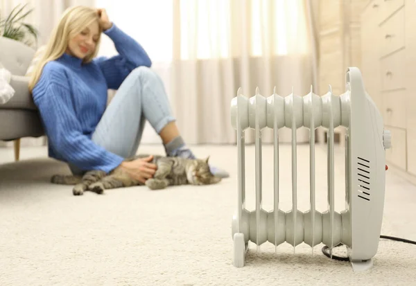 Calentador eléctrico y mujer joven borrosa con lindo gato tabby en el fondo. Espacio para texto — Foto de Stock