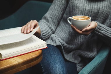 Elinde kahve fincanı olan bir kadın evde kitap okuyor.