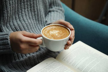 Elinde kahve fincanı olan bir kadın evde kitap okuyor.