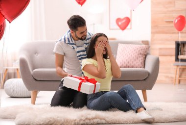 Genç adam oturma odasında kalp şeklinde balonlarla süslenmiş kız arkadaşına hediye sunuyor. Sevgililer Günü kutlaması