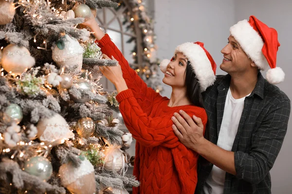 Happy young couple in Santa hats decorating Christmas tree toget — Stock Photo, Image