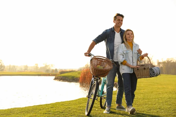 阳光明媚的日子 一对年轻夫妇在湖边骑着自行车和野餐篮 — 图库照片