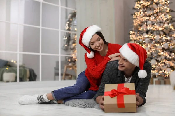 Happy young couple in Santa hats with Christmas gift at home — Stock Photo, Image