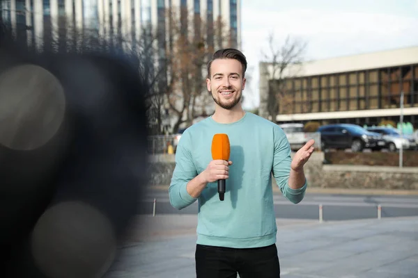 Young male journalist with microphone working on city street